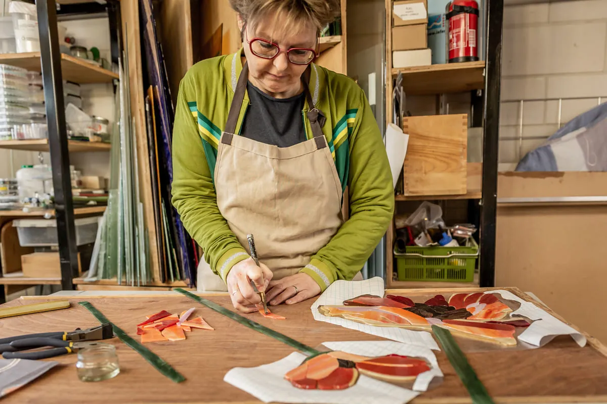 Liesbeth Kocken met schort aan de werktafel in haar atelier, bezig met gekleurd glas