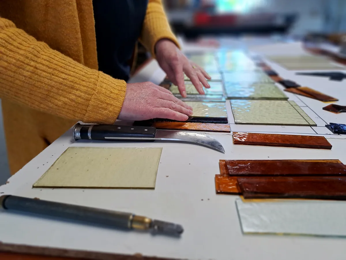 Handen leggen gesneden stroken amberkleurig glas klaar op de werktafel, met glassnijder en tang ernaast